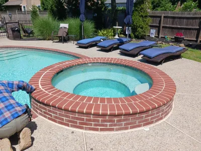 A raised circular brick spa and adjacent swimming pool are surrounded by an aggregate patio and lounge chairs in a sunny backyard setting while a worker inspects the edge.