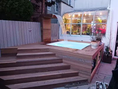 A modern wooden deck surrounds an illuminated spa featuring a cascading waterfall, set against a backdrop of a plant-filled conservatory at dusk.