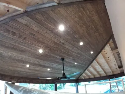 A curved ceiling made of dark wood planks features recessed lighting and a central ceiling fan, viewed alongside unfinished construction elements.