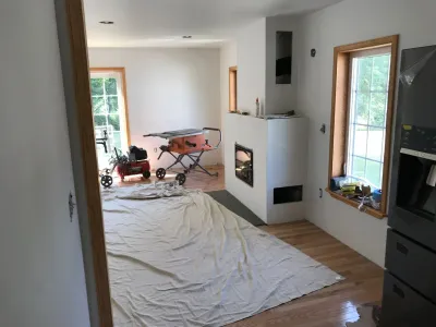 A residential room undergoing renovation features a new modern fireplace, hardwood floors, natural light windows, and construction tools on a drop cloth.